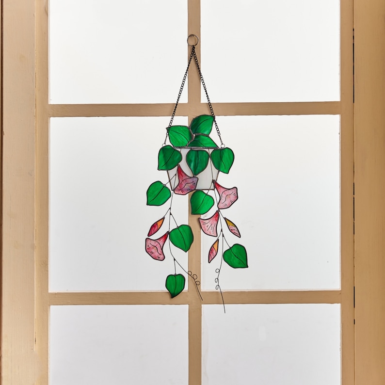 Peut inclure: Une suspension en verre color&eacute; repr&eacute;sentant une plante en pot avec des fleurs roses et des feuilles vertes. La plante est suspendue &agrave; une cha&icirc;ne noire.