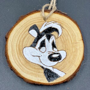 Puede incluir: Un adorno redondo de madera con un dibujo de mofeta de dibujos animados en blanco y negro pintado en él. La mofeta está mirando hacia la derecha y tiene una cara blanca con ojos negros y una nariz negra.
