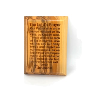May include: A wooden plaque with the Lord's Prayer engraved in brown text. The plaque is made of olive wood and has a natural wood grain. The text is centered on the plaque and is surrounded by a border. The plaque is a religious item that can be used for prayer or meditation.