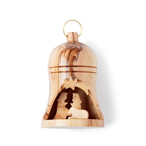 May include: A wooden bell-shaped Christmas ornament with a carved nativity scene. It has a gold-coloured metal loop for hanging. The wood grain is visible, with light and dark brown tones. The ornament is approximately 10 cm tall.
