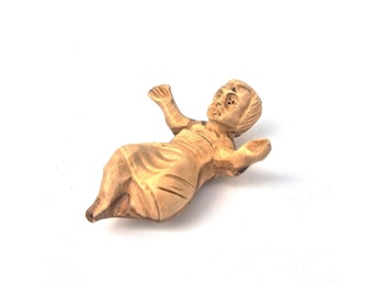 May include: A small, hand-carved wooden figurine depicting a reclining figure with arms outstretched. The sculpture features natural light brown wood grain patterns, visible facial features, and carved drapery folds across the body.