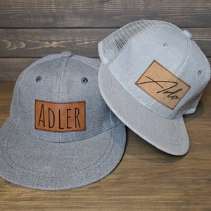 May include: Two gray baseball caps with brown leather patches. One patch says "Adler" and the other says "Arlo".
