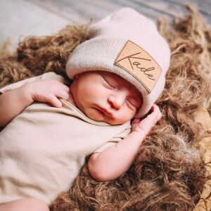 May include: A newborn baby wearing a beige knitted beanie with a brown leather patch that says "Kade" is sleeping in a brown fluffy basket.
