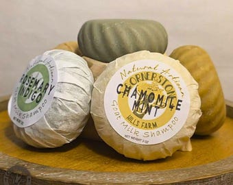 May include: A collection of round, ridged goat milk shampoo bars. Two bars are wrapped in paper with labels reading Rosemary Indigo and Chamomile Mint, while two unwrapped bars in olive green and tan are visible behind them. Each bar weighs 3 oz.