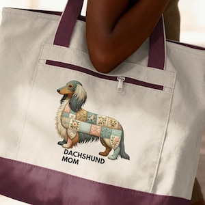 May include: Cream-colored tote bag with a burgundy base and straps. The bag features a dachshund illustration with a patchwork design and the text "DACHSHUND MOM". The bag has a zippered pocket and is carried over the shoulder.