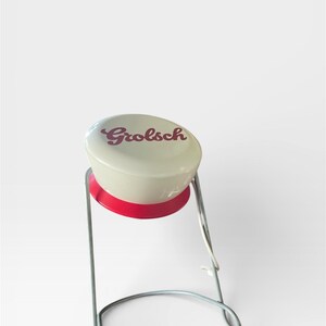 Op de afbeelding: Een Grolsch bierflesstopper met een witte bovenkant en rode basis, ondersteund door een zilverkleurig metalen frame. Het woord "Grolsch" staat in rode letters op de bovenkant.