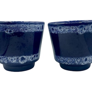 Può includere: Due fioriere in ceramica blu navy con una fascia bianca e strutturata intorno alla parte superiore. Le fioriere hanno una forma svasata e una base scura. Queste fioriere decorative sono adatte per uso interno o esterno.
