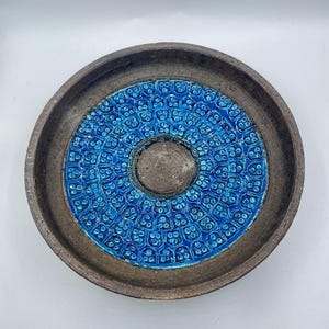 May include: A round ceramic plate with a dark brown rim and a bright blue centre. The centre has a repeating pattern of small, flower-like shapes. The plate has a small, raised centre and is set against a white background.