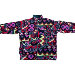 Peut inclure: Un pull en polaire coloré avec un motif géométrique dans les tons de rose, violet, rouge, vert et gris. Le haut à manches longues a un col montant et une fermeture à quart de fermeture éclair. Le nom de la marque est brodé sur la poitrine.