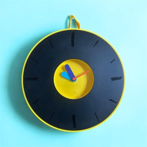 Pode incluir: Um relógio redondo amarelo com números pretos e um mostrador amarelo com uma mão azul, vermelha e laranja. O relógio está pendurado em uma parede azul claro.