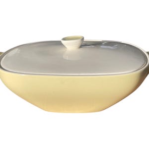 Könnte beinhalten: Ovale Servierschüssel mit Deckel. Die Schüssel ist hellgelb, der Deckel weiß, mit einem kleinen Griff. Sie hat zwei Griffe an den Seiten. Vintage Küchenartikel.