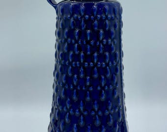 Może przedstawiać: Wazon ceramiczny w kolorze kobaltowym z fakturowaną powierzchnią i uchwytem. Wazon ma zaokrąglone dno i lekko rozszerzającą się górę.