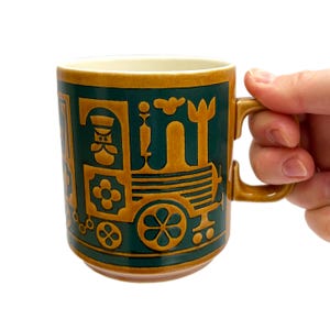 May include: A ceramic mug with a train design. The mug features a dark green background with a raised, tan-colored train illustration. The handle and rim are also tan. The mug is held by a hand.