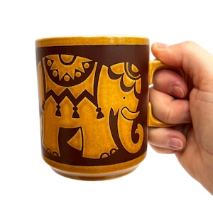 May include: A brown ceramic mug with a mustard yellow elephant design. The elephant is decorated with intricate patterns. The mug has a square handle and a brown rim. The mug is held by a hand.