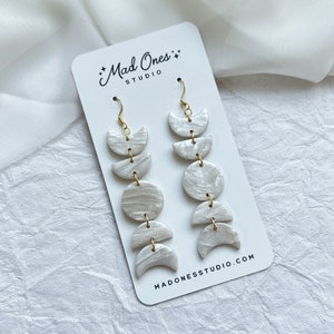 May include: White moon phase earrings with gold hooks. The earrings are made of polymer clay and have a marbled effect. The earrings are hanging on a white card with the text "Mad Ones Studio" and "Madonessstudio.com".