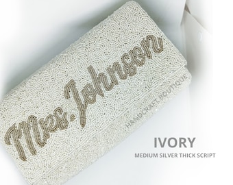 May include: Ivory beaded clutch with the text "Mrs. Johnson" in silver script.