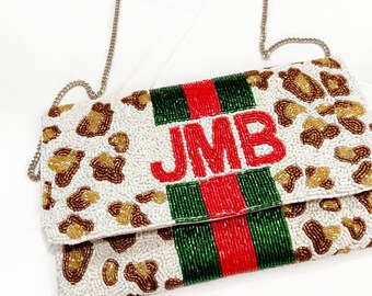 Könnte beinhalten: Eine weiße Perlen-Clutch mit Leopardenmuster. Die Tasche hat ein grün-rot-weiß gestreiftes Muster mit den Initialen "JMB" in roten Perlen. Die Tasche hat einen silbernen Kettenriemen.