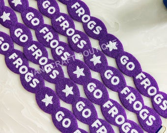 May include: Purple beaded straps with the text "GO FROGS" in white, interspersed with white star shapes. The straps have gold-colored clasps at the ends.