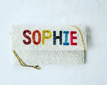 Könnte beinhalten: Weiße Perlenclutch mit goldener Kettenriemen. Die Clutch trägt den Namen "SOPHIE" in bunten Perlen.