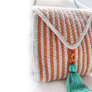 Op de afbeelding: Een gestreepte gehaakte tas met een lange kwast. De tas is gemaakt van beige en oranje garen en heeft een klep sluiting. De kwast is helder turquoise.