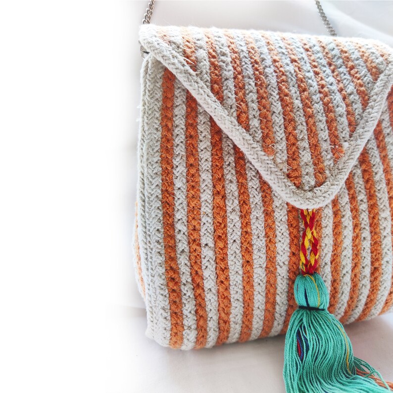 Op de afbeelding: Een gestreepte gehaakte tas met een lange kwast. De tas is gemaakt van beige en oranje garen en heeft een klep sluiting. De kwast is helder turquoise.