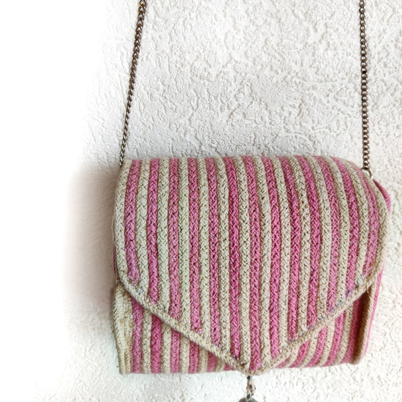 Op de afbeelding: Een gestreepte, gehaakte tas in roze en beige met een zilveren kettingband en een zilveren hanger die aan de onderkant hangt.