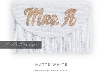 Könnte beinhalten: Weiße Perlenclutch mit einer dicken Schrift in Champagnergold, die "Mrs. A" lautet. Die Clutch hat einen Fransenrand.