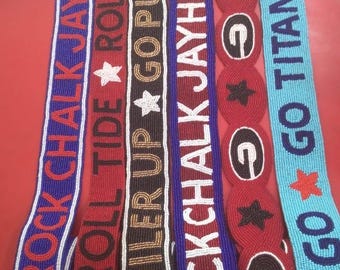 May include: Six beaded straps featuring various sports team slogans and logos. The straps include text such as ROCK CHALK JAYHAWK, ROLL TIDE, BOILER UP, and GO TITANS GO, along with star and letter G symbols in blue, red, black, white, and light blue.