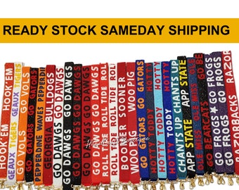 May include: A collection of 24 colorful, glittery, fabric keychains with various college team logos and names, including "Hook 'Em", "Geaux Tigers", "Go Vols", "Go Gators", "Pepperdine Waves", "Georgia Bulldogs", "Go Dawgs", "Roll Tide", "Boomer Sooner", "Woo Pig", "Hotty Toddy", "Chants Up", "App State", "Ole Miss", "Go Bearcats", "Go Frogs", and "Razorbacks".