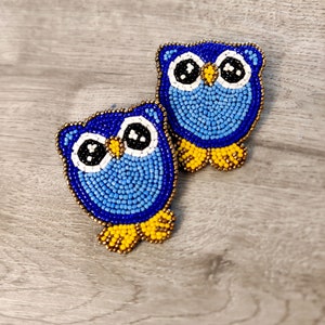 Könnte beinhalten: Ein Paar blaue und gelbe Perlenohrringe in Form von Eulen. Die Eulen haben weiße Augen und einen gelben Schnabel.