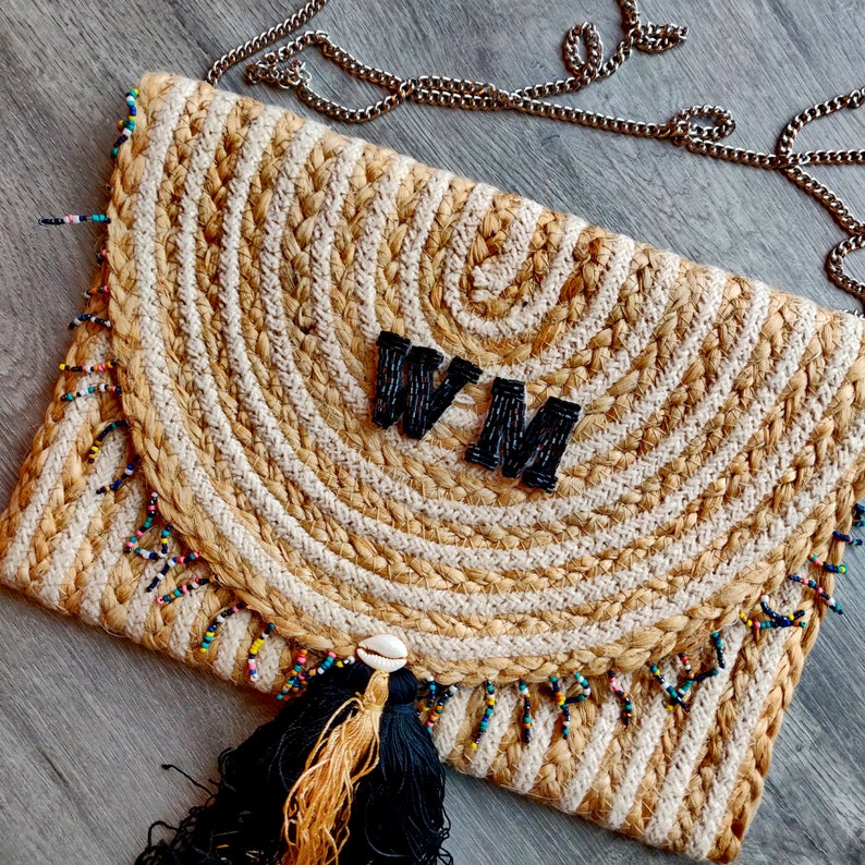 May include: A beige and white woven clutch bag with a black tassel and beaded fringe. The bag has a black leather flap with the initials "WM" in black.