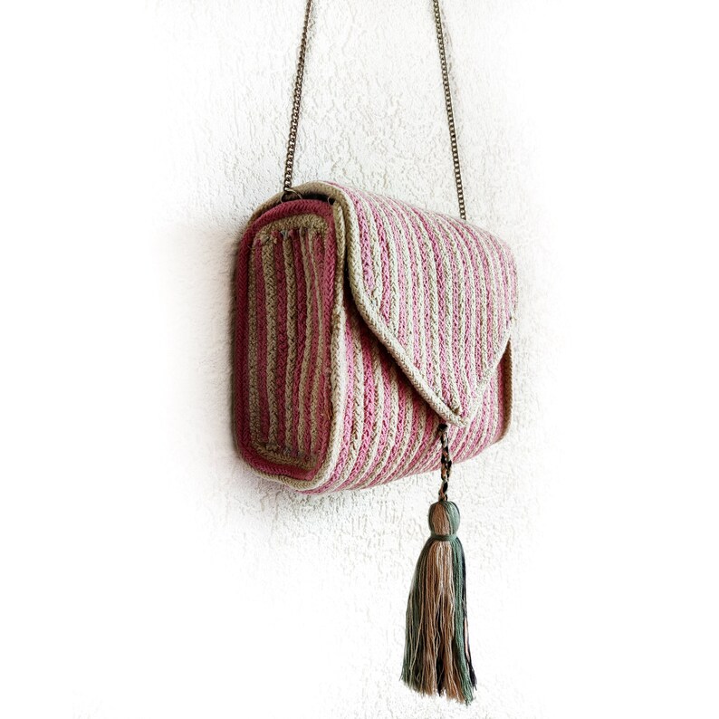 Op de afbeelding: Een gestreepte, gehaakte schoudertas in roze en beige met een kwast. De tas heeft een kettingband en een klep sluiting.