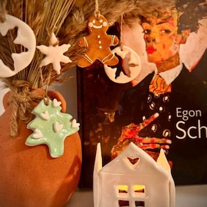 May include: A terracotta vase containing dried grasses and hanging ceramic ornaments including a gingerbread man, a white snowflake, a white star, and a light green Christmas tree with white hearts, next to a white ceramic house-shaped candle holder.