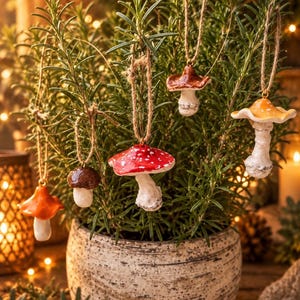 May include: Five ceramic mushroom ornaments hanging from a rosemary plant. The mushrooms feature various cap colors including bright red with white spots, orange, brown, and tan, each suspended by a thin brown twine loop for decorative display.