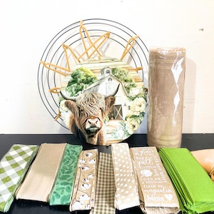 May include: A farmhouse-themed wreath with a Highland cow and barn scene, surrounded by a black metal frame and gold accents. Includes a roll of burlap ribbon, and various patterned ribbons in green, tan, and white. Some ribbons have text.