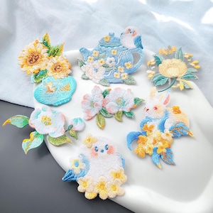 May include: A collection of seven embroidered fabric patches featuring various designs including yellow sunflowers in a blue vase, a blue teapot with a bird, a white flower, a rabbit with yellow flowers, and a small bird surrounded by yellow blossoms.