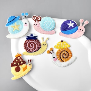 May include: A collection of six embroidered snail patches featuring various designs. The snails have colourful shells, some wearing hats like a blue cap, a sailor hat, or a yellow umbrella. Each snail has distinct facial features and antennae.