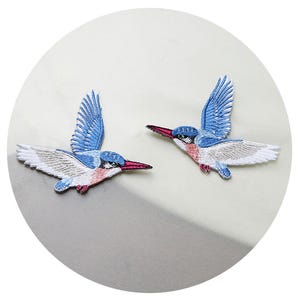 May include: Two blue and white embroidered bird patches with red beaks. The birds are in a flying position with their wings spread out.