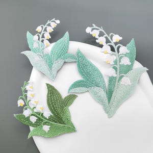 Op de afbeelding: Drie geborduurde patches met lelietje-van-dalen bloemen. Elke patch toont witte klokvormige bloesems met gele harten en groene bladeren. Twee patches zijn in licht mintgroen en één is in een donkerdere tint groen uitgevoerd.
