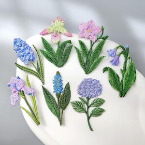 May include: A collection of seven embroidered flower patches featuring various botanical designs. The set includes blue hyacinths, a pink and green orchid, a purple flower, blue bell-shaped blossoms, a purple iris, a blue spike flower, and a hydrangea.