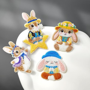 Peut inclure: Quatre écussons brodés représentant des lapins marron et blancs. Un lapin porte un chapeau jaune sur une étoile jaune. Un autre porte une couronne de fleurs. Un troisième porte un chapeau bleu et une écharpe violette. Le quatrième porte un chapeau bleu.