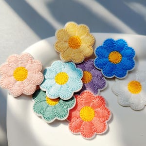 May include: A collection of small, embroidered flower patches in various colors including yellow, blue, light blue, pink, green, purple, orange, and white. Each patch features a circular yellow center surrounded by eight rounded petals.