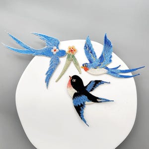 May include: Three embroidered bird patches featuring two blue swallows and one black and blue swallow. One blue swallow holds a small orange flower with green leaves in its beak. The embroidery uses detailed thread work to show wing feathers.