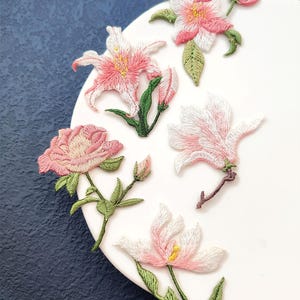 Puede incluir: Parches florales bordados en tonos rosa, blanco y verde. Los parches presentan varios diseños florales, incluyendo lirios y rosas, con costuras detalladas. Los parches están dispuestos sobre una superficie blanca sobre un fondo azul oscuro.