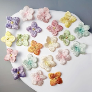 May include: A collection of embroidered flower appliques in various pastel colors, including pink, purple, yellow, and green. Each flower has a detailed, textured appearance and a small, golden center. The appliques are arranged on a white surface.