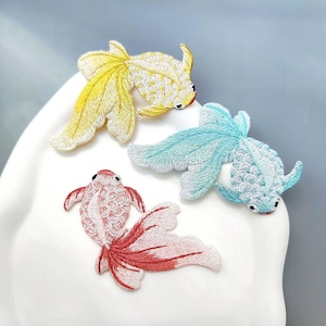 May include: Three embroidered goldfish patches in yellow, blue, and red. The patches are detailed with intricate stitching, showcasing the fish's fins and scales. The patches are placed on a white surface against a light blue background.