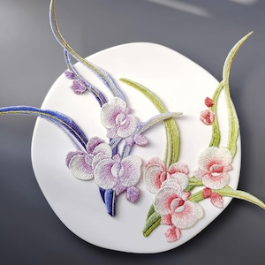 Puede incluir: Obra de arte floral bordada sobre una base blanca y circular. El diseño presenta flores detalladas similares a orquídeas en tonos morados, rosas y blancos, con acentos verdes y azules. La obra de arte está sobre un fondo gris.