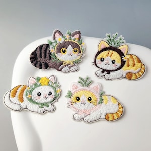 May include: Four embroidered patches of cartoon cats, each wearing a floral or leafy crown. The cats are in a lying down position and have different colour patterns, including brown, orange, and white, with embroidered details.