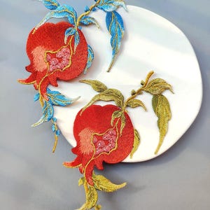 May include: Two embroidered pomegranate appliques. The pomegranates are red with pink and gold detailing, and the leaves are blue and gold. The appliques are on a white plate, with a grey background. Ideal for fabric crafts.