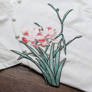 Puede incluir: Parche bordado para planchar con un diseño floral con flores rosas y blancas y hojas verdes.